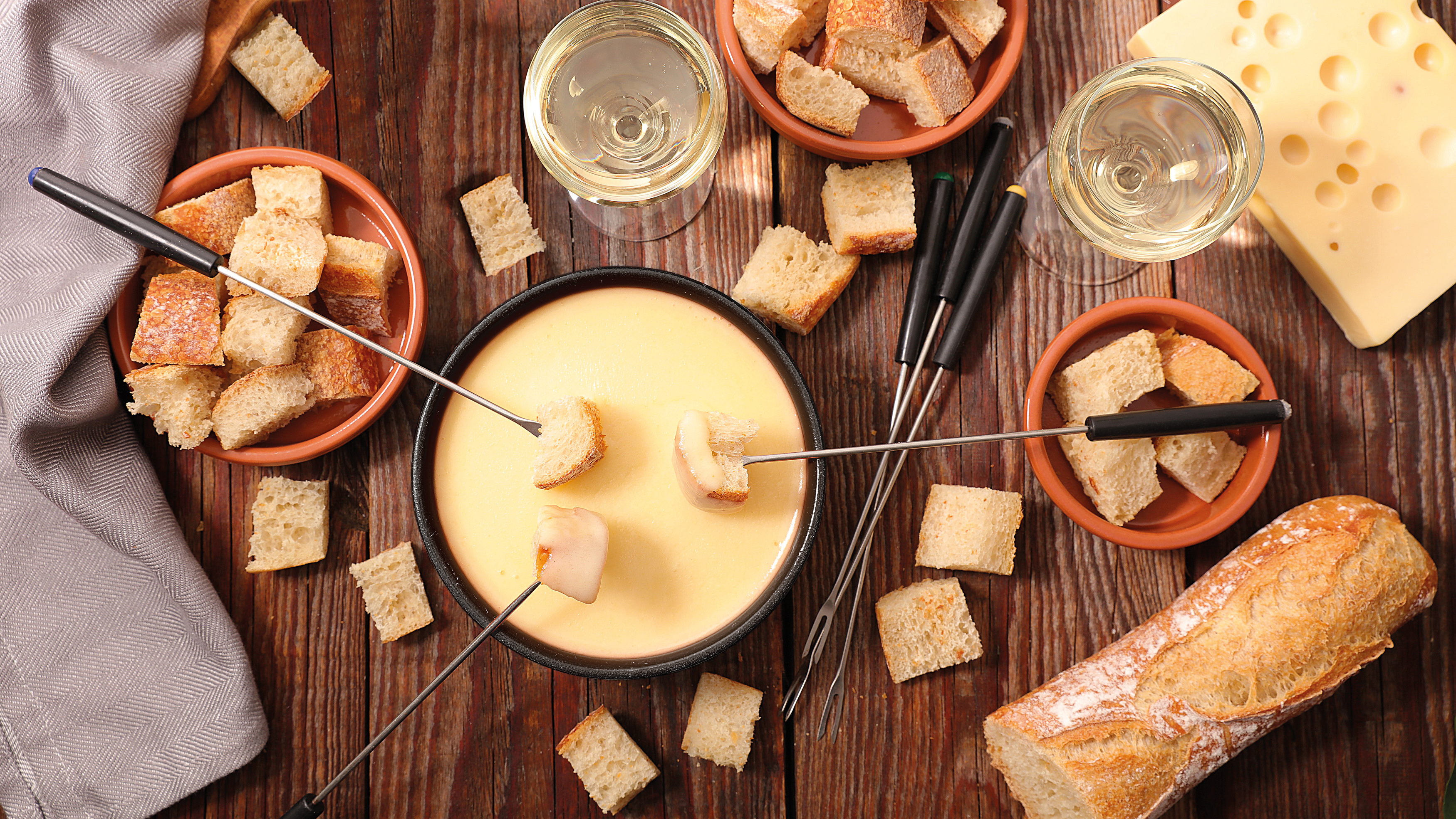 Ein Fondue-Set mit Brot, Käse, Wein und Fonduegabeln auf einem Holztisch, Draufsicht.