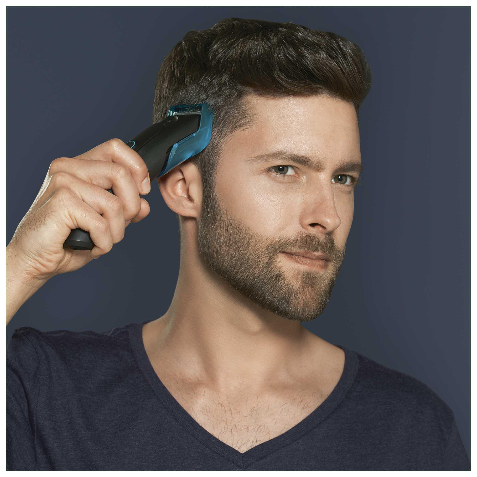 Ein Mann mit Bart benutzt einen schwarz-blauen Haarschneider, vor blauem Hintergrund.