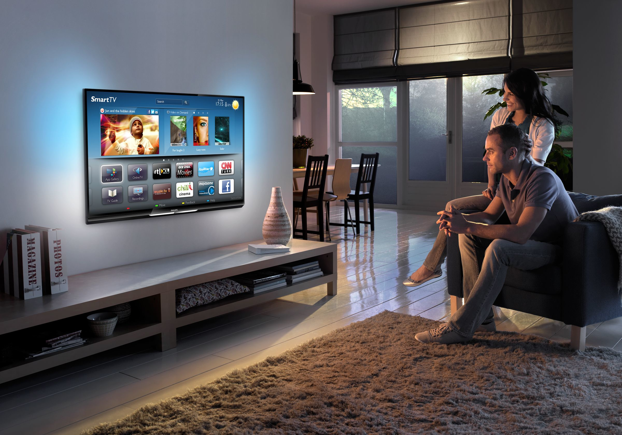 Salón con TV. Hombre y mujer sentados. Pantalla TV mostrando apps. Libros y florero.