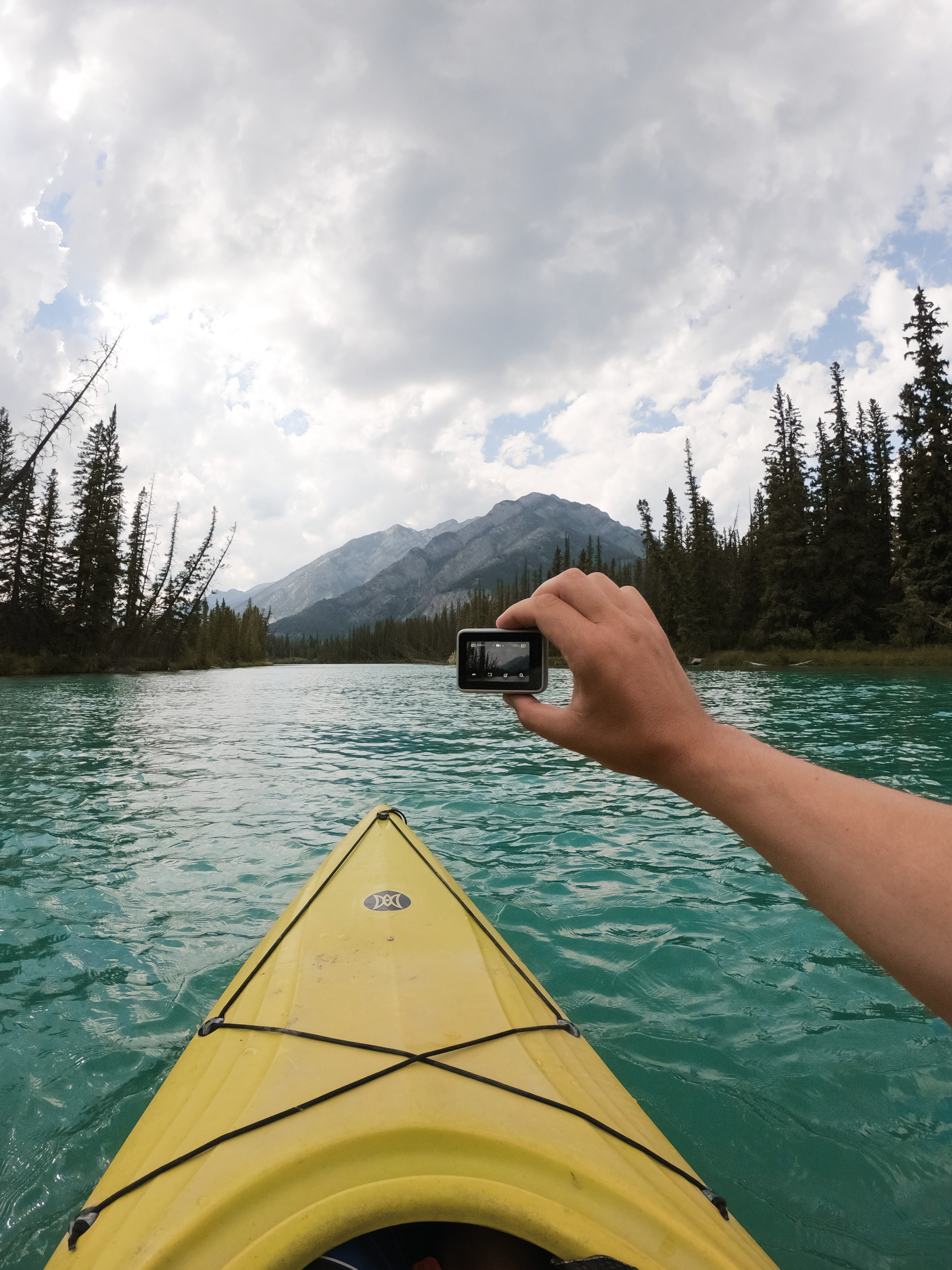 Person Kajakfahren auf einem teal-farbenen See, hält eine Kamera, Berge im Hintergrund.