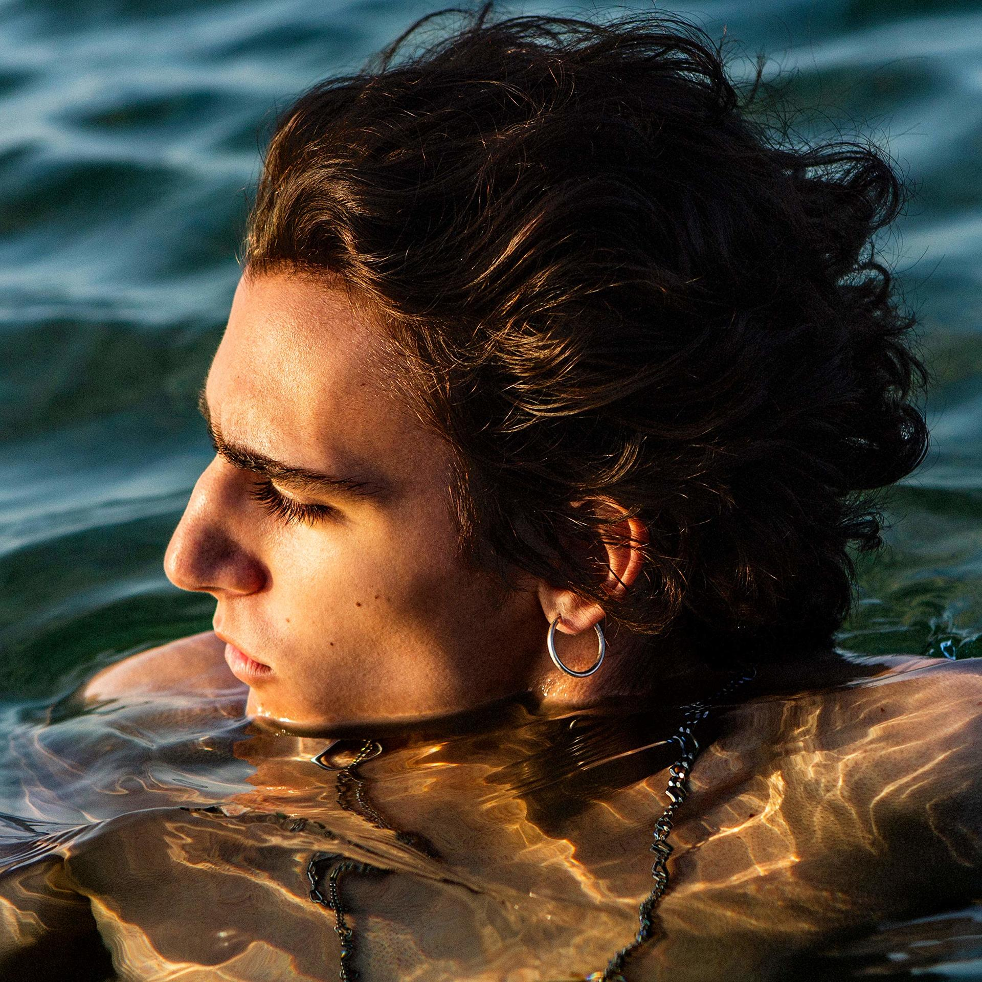 Homme dans l'eau, regardant ailleurs, l'eau reflète la lumière du soleil. Il porte une chaîne et une boucle d'oreille.
