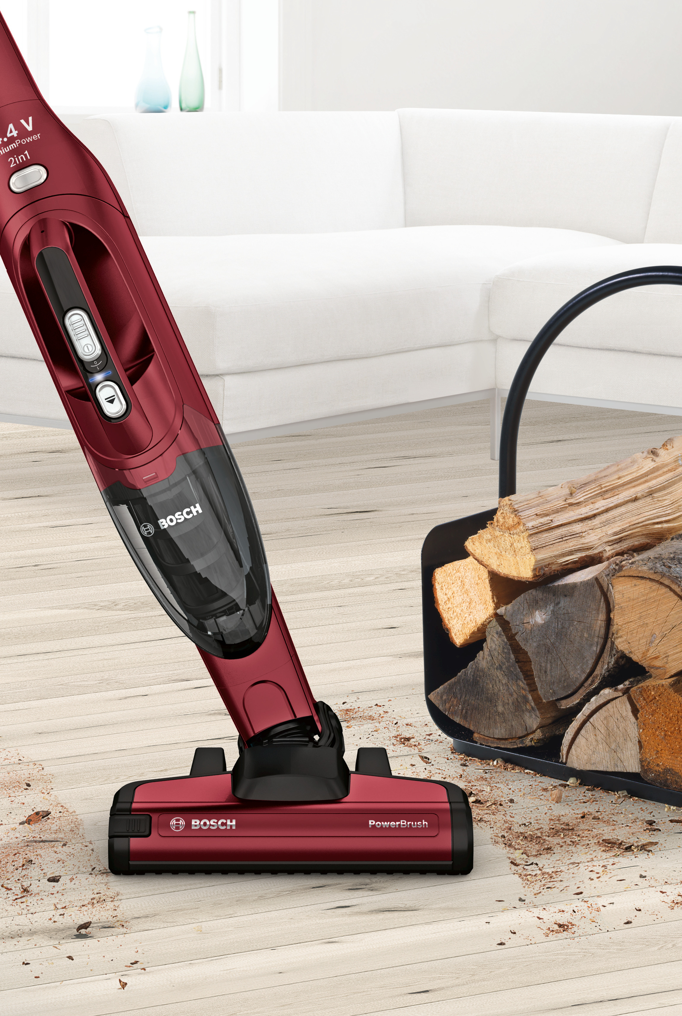 Ein roter Bosch PowerBrush Staubsauger reinigt einen Holzboden. Holzscheite liegen in einem schwarzen Halter. Eine weiße Couch im Hintergrund.