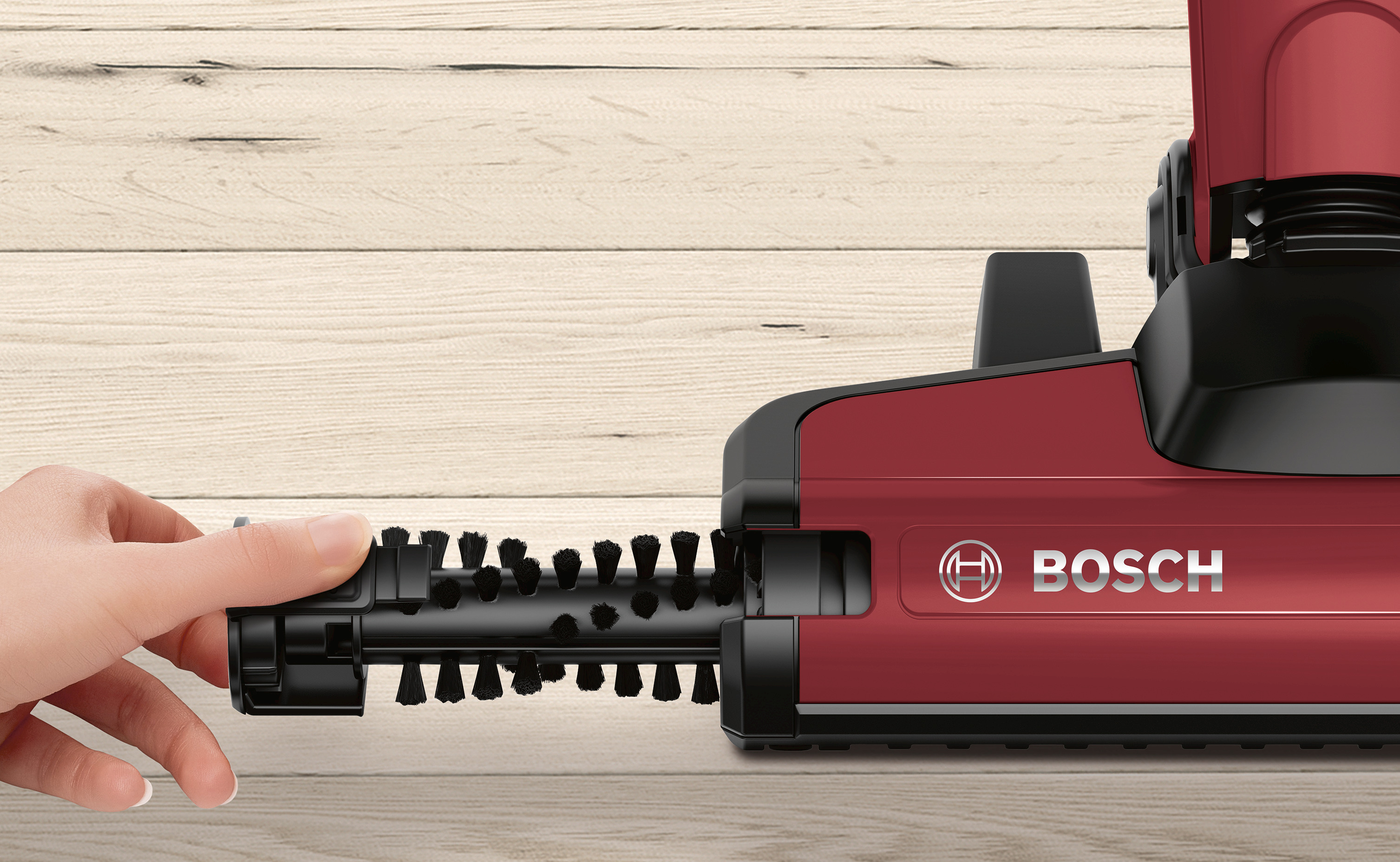 Hand hält Vakuum-Bürste. Roter Staubsaugerkörper. Heller Holzuntergrund. Bosch-Logo sichtbar.