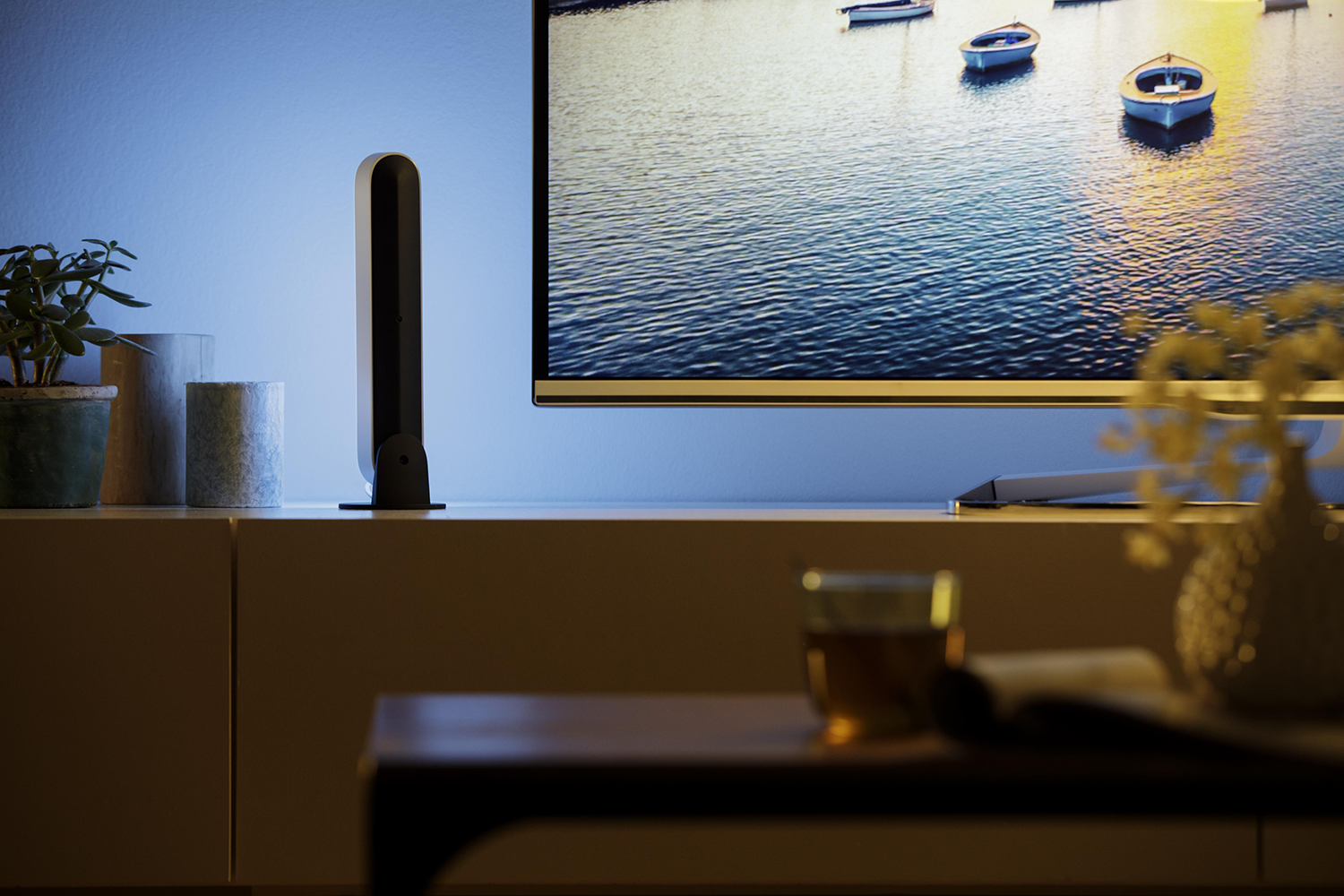 An illuminated light bar is on a white cabinet. A tv is in the background. A cup and vase are on the cabinet.