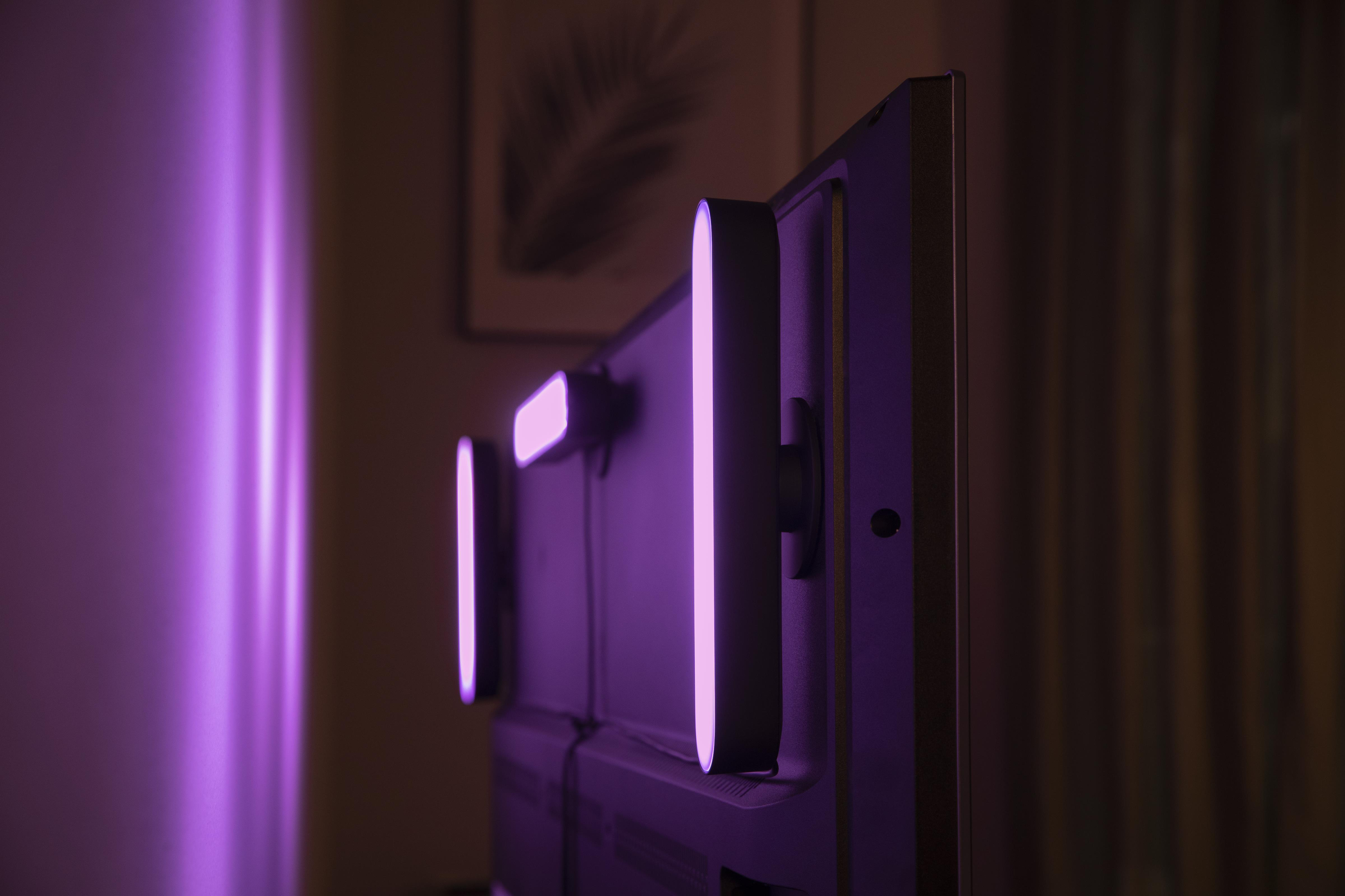 Three purple lights on a dark TV against a purple-lit wall. Side view.
