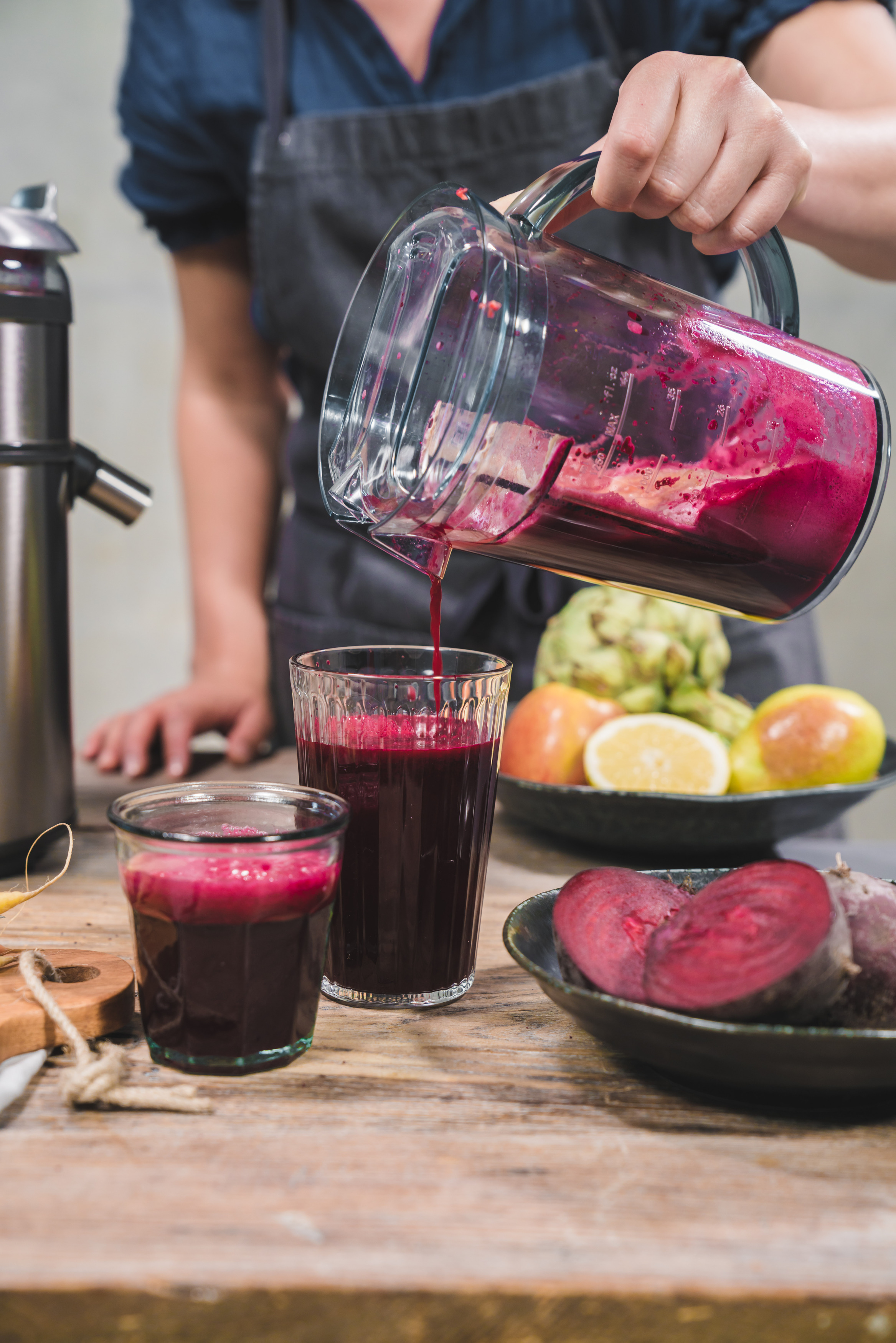 Eine Person gießt Saft in zwei Gläser. Rote Bete, Zitronen, Birnen und ein Entsafter stehen auf dem Holztisch.