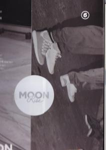 Een grijswaardenafbeelding met voeten in witte sneakers in de buurt van een rond 'MOON RISE' logo.