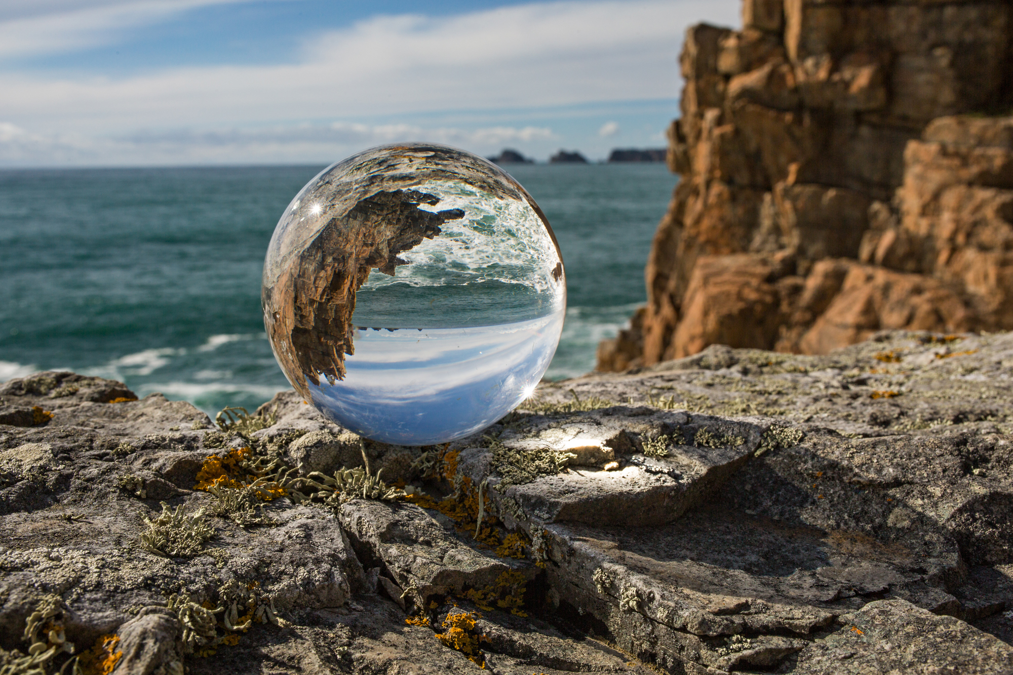 Glaskugel auf einer felsigen Klippe. Die Kugel spiegelt Meer, Himmel und Klippen wider. Sonnenschein.