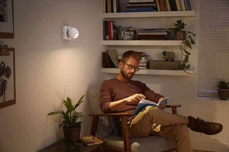Un hombre leyendo un libro bajo una lámpara de pared, sentado en un sillón. Estantería al fondo.