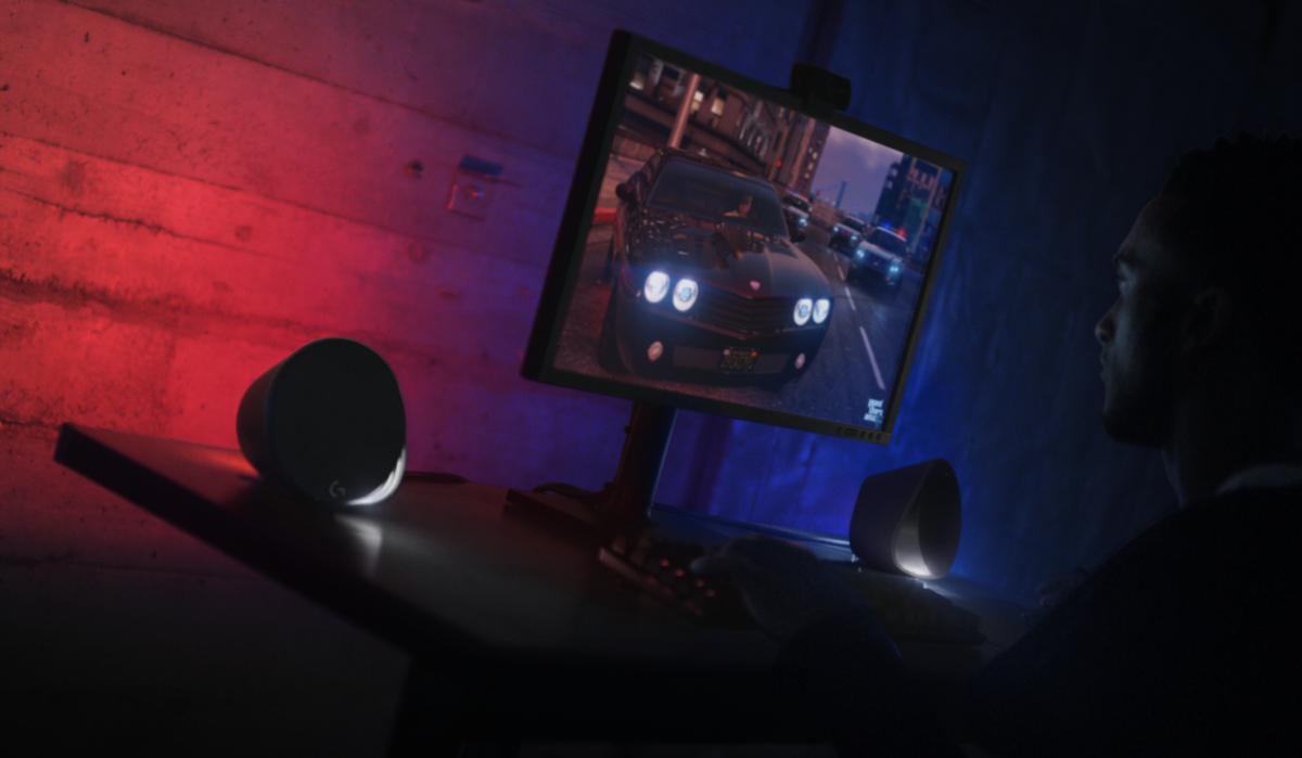 Una persona está jugando en una computadora. La pantalla muestra un coche en una ciudad. Luces rojas y azules en el fondo.