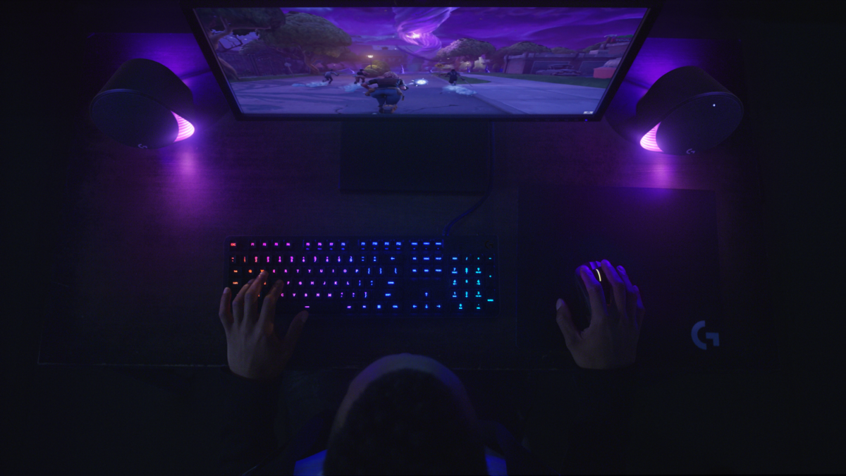 Vista cenital de persona jugando con teclado, ratón, monitor y altavoces iluminados en morado.