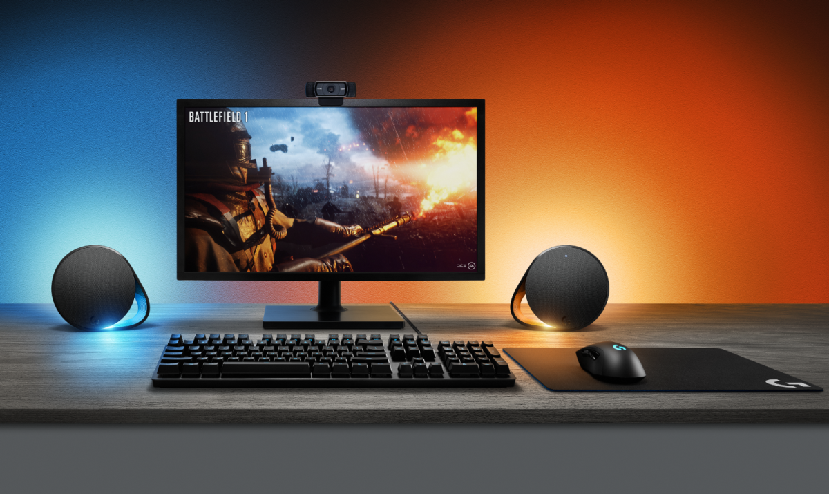 Configuración de juego con monitor mostrando Battlefield 1, teclado, ratón, altavoces y webcam.
