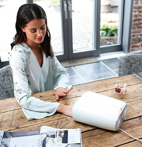 Mujer haciéndose las uñas en una mesa. Secador de uñas blanco. Botellas de esmalte de uñas. Luz natural y mesa de madera.