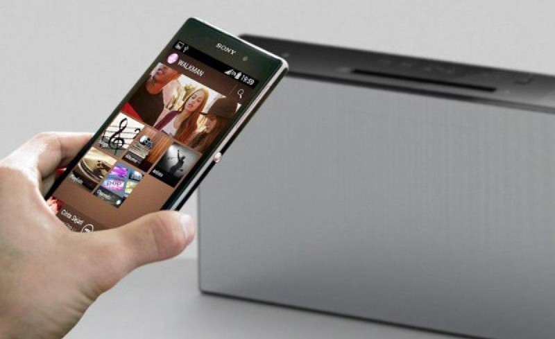 Una mano sosteniendo un smartphone que muestra una aplicación de música, junto a un altavoz gris.