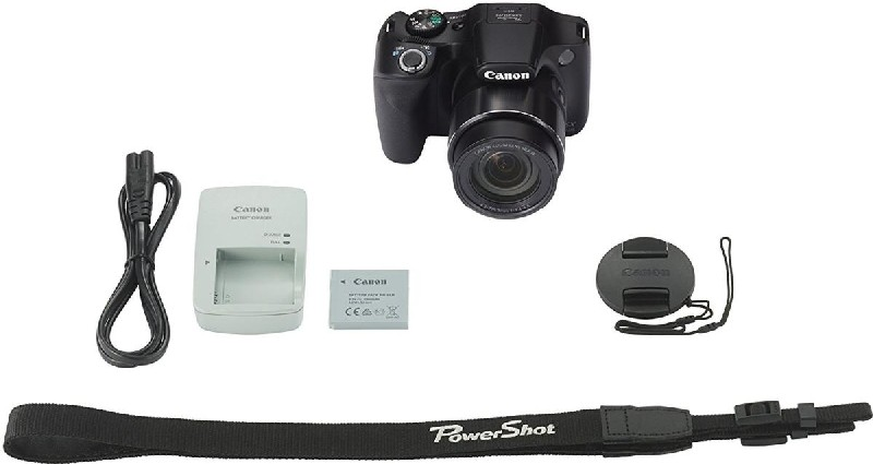 Cámara Canon, cargador, batería, tapa del objetivo, correa, todo negro, sobre fondo blanco.