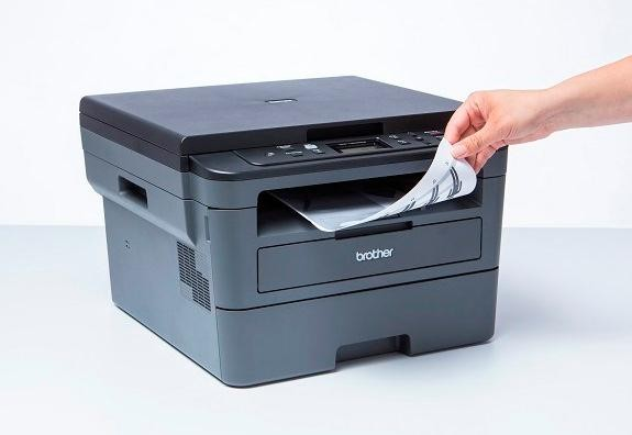 Una impresora gris y negra con una mano quitando un papel impreso. Fondo blanco.