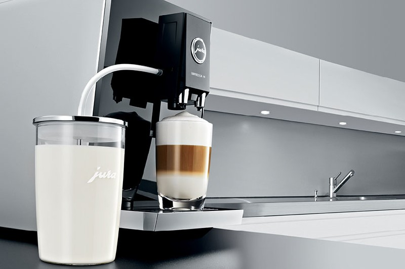 Máquina de espresso con recipiente de leche de vidrio y un vaso de latte frente a una encimera de cocina.