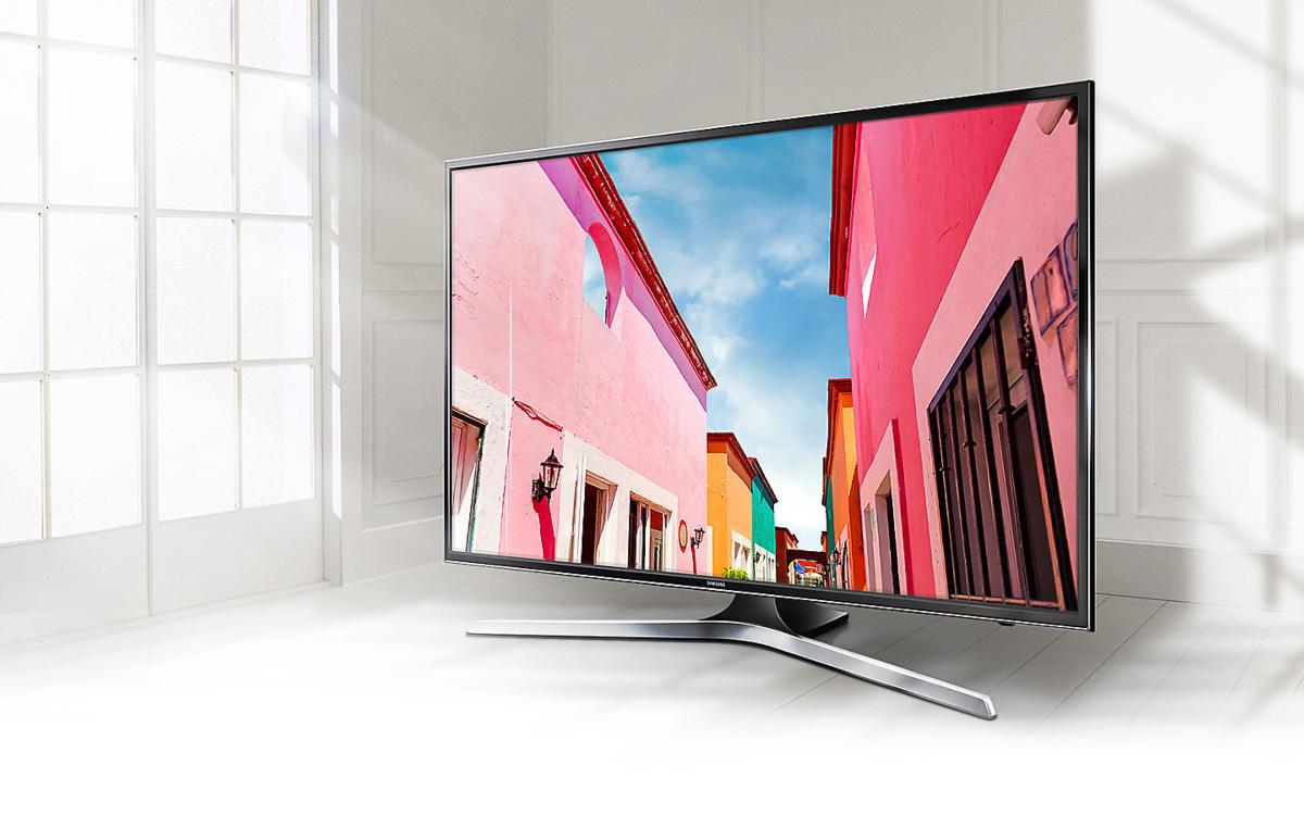 Un televisor Samsung que muestra una escena callejera colorida. Tiene un soporte plateado, en un suelo blanco.