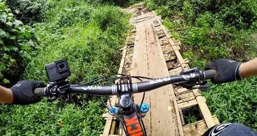 Vista en primera persona de un ciclista de montaña en un puente de madera sobre una zona verde.