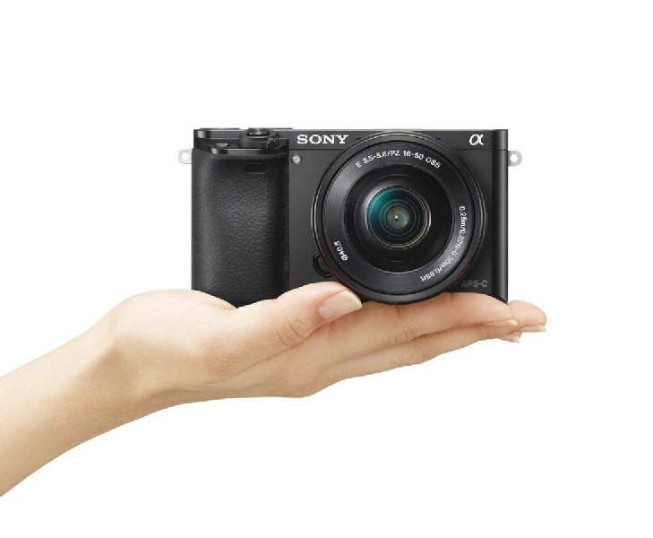 La mano de una persona sosteniendo una cámara Sony, negra, con una lente, contra un fondo blanco.