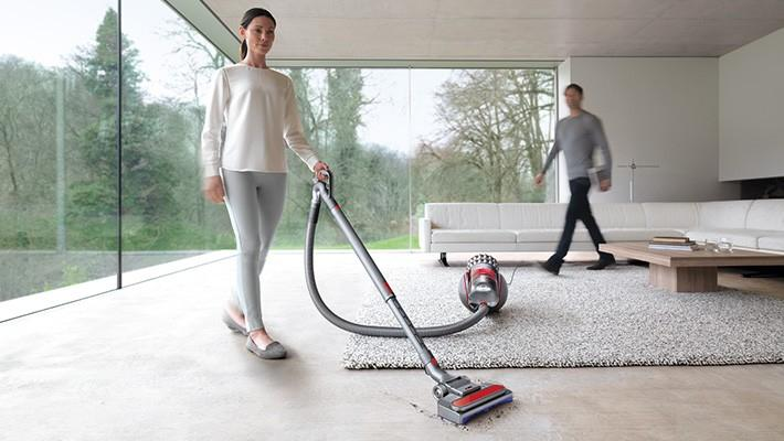 Una mujer aspira una alfombra mientras un hombre pasa por una sala de estar moderna, sofá blanco visible.