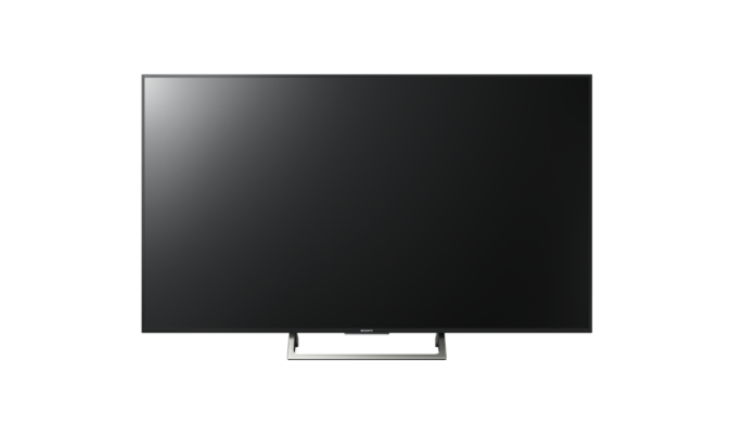 Televisor de pantalla plana grande y negro sobre un soporte, sobre un fondo blanco. La pantalla está apagada.