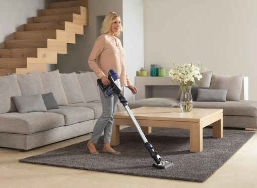 Una mujer aspirando una alfombra gris con una aspiradora inalámbrica. Sofá, mesa y escaleras al fondo.