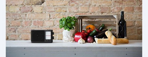 Radio negro sobre una superficie blanca, con verduras y vino de fondo.