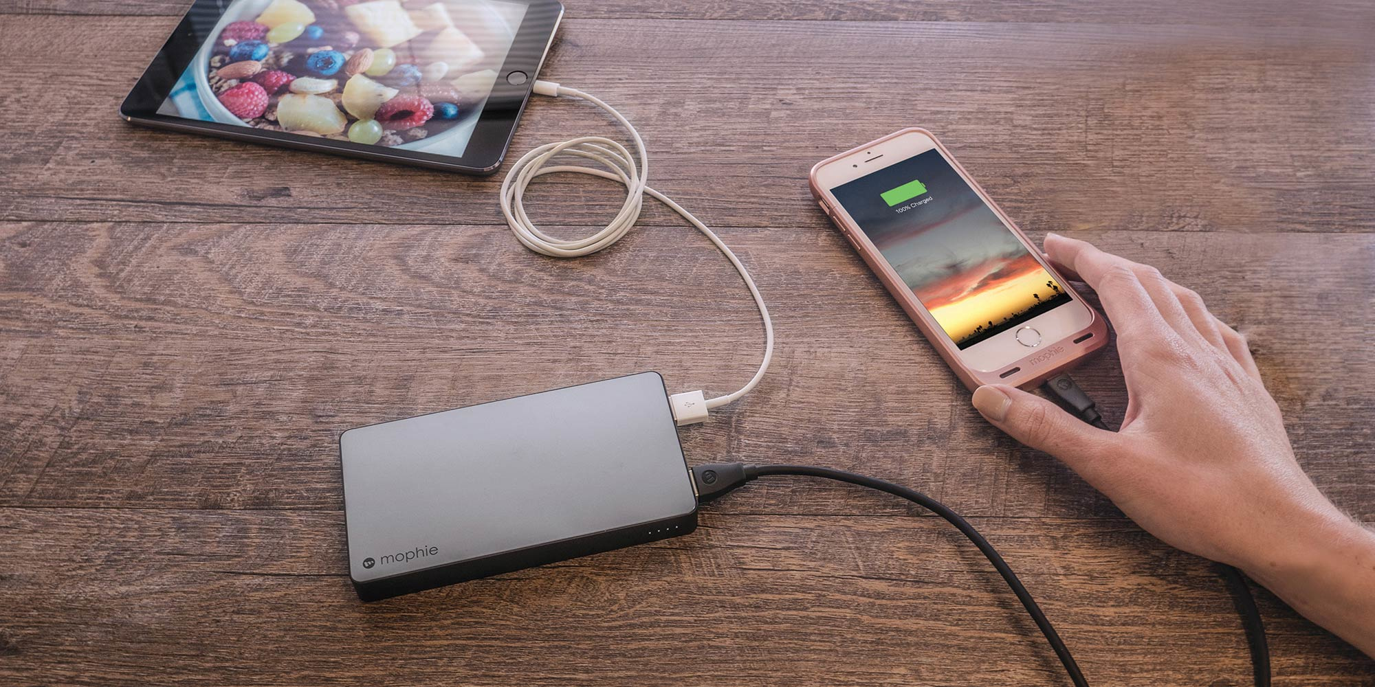 Una persona carga un teléfono y una tableta. Un banco de energía está sobre una mesa de madera. El teléfono muestra 100% cargado.