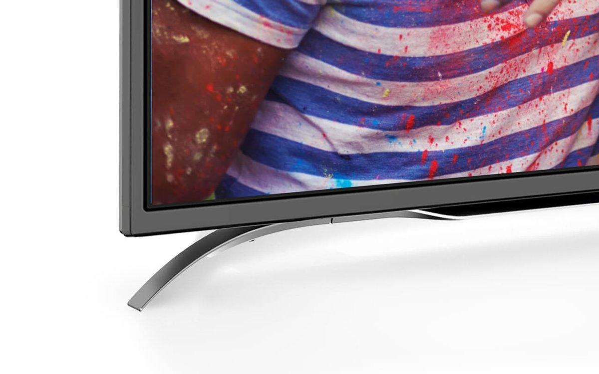Un televisor negro con un soporte plateado curvo. Una persona es parcialmente visible en el fondo con pintura.