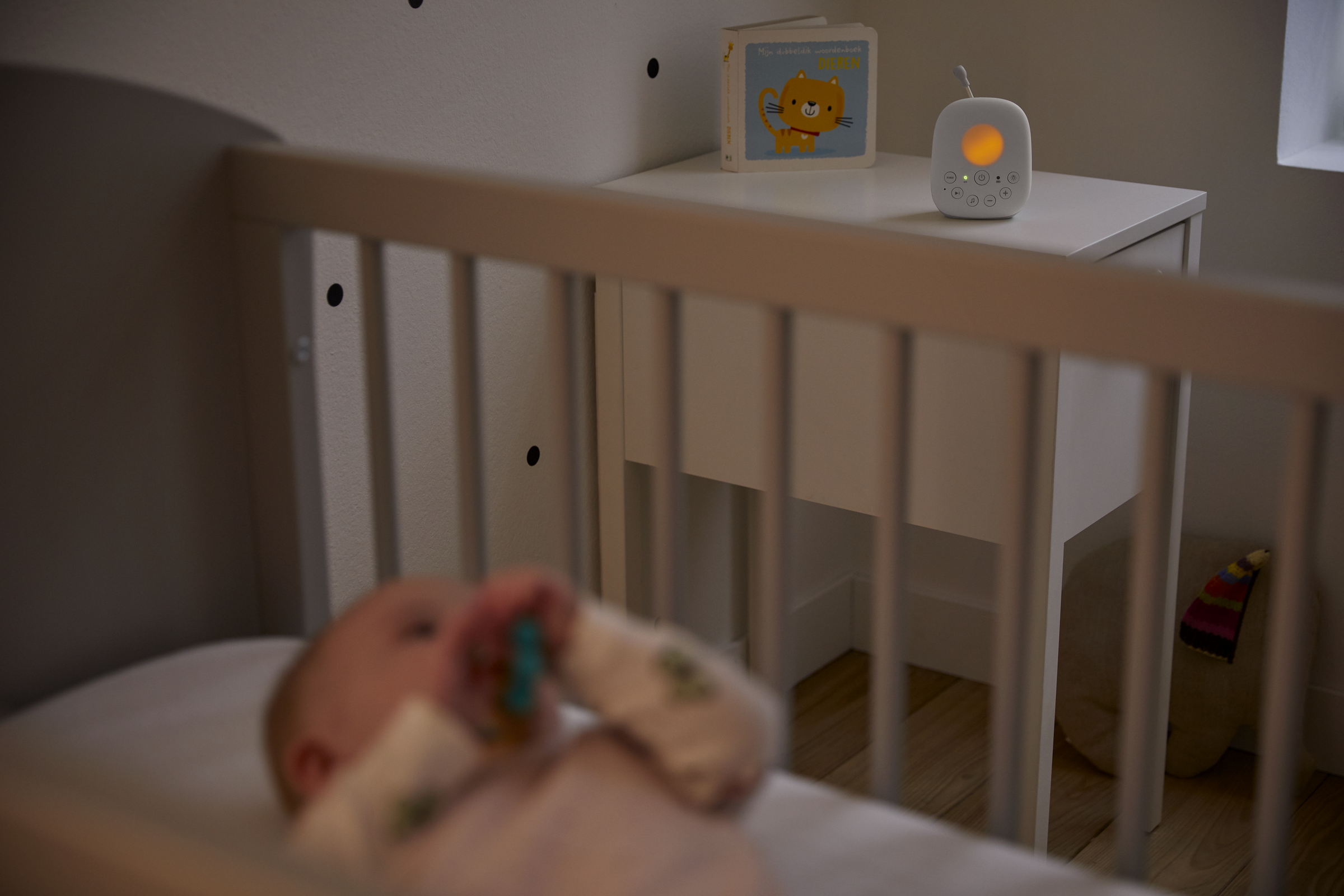 A baby in a crib with a pacifier, a baby monitor, and a book on a nightstand.