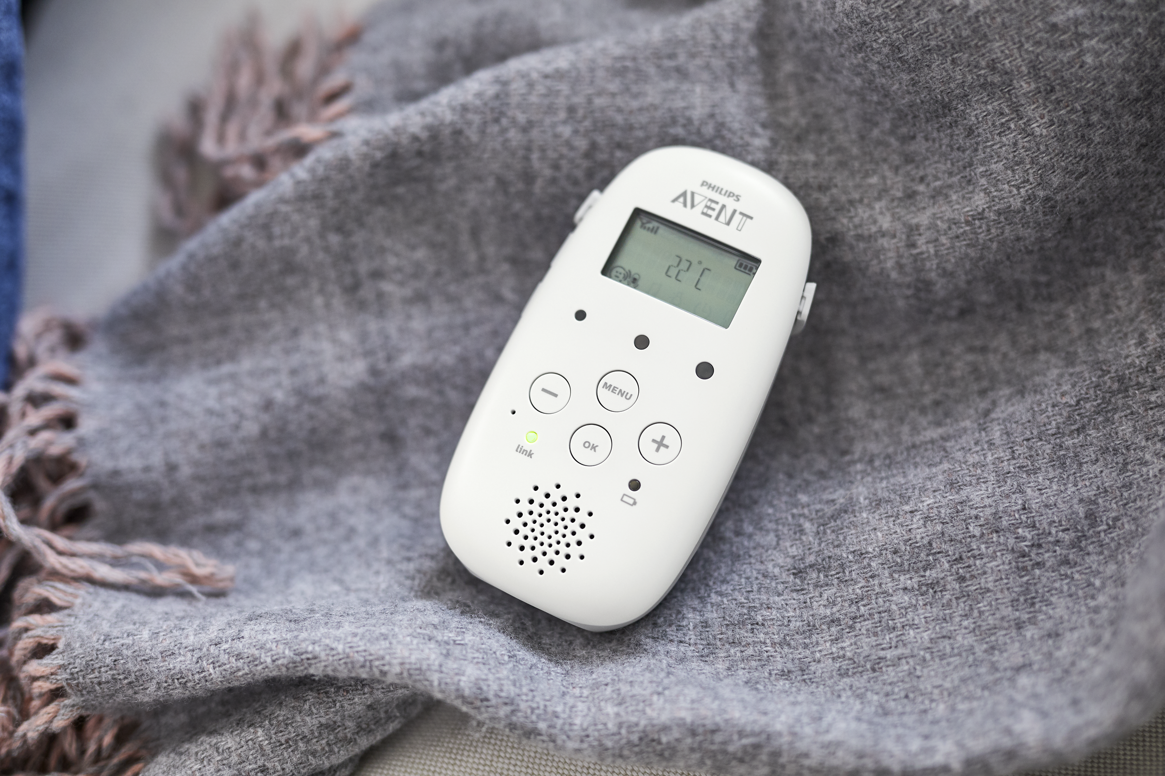 A white baby monitor with a screen, buttons, and a speaker, lying on a blanket.