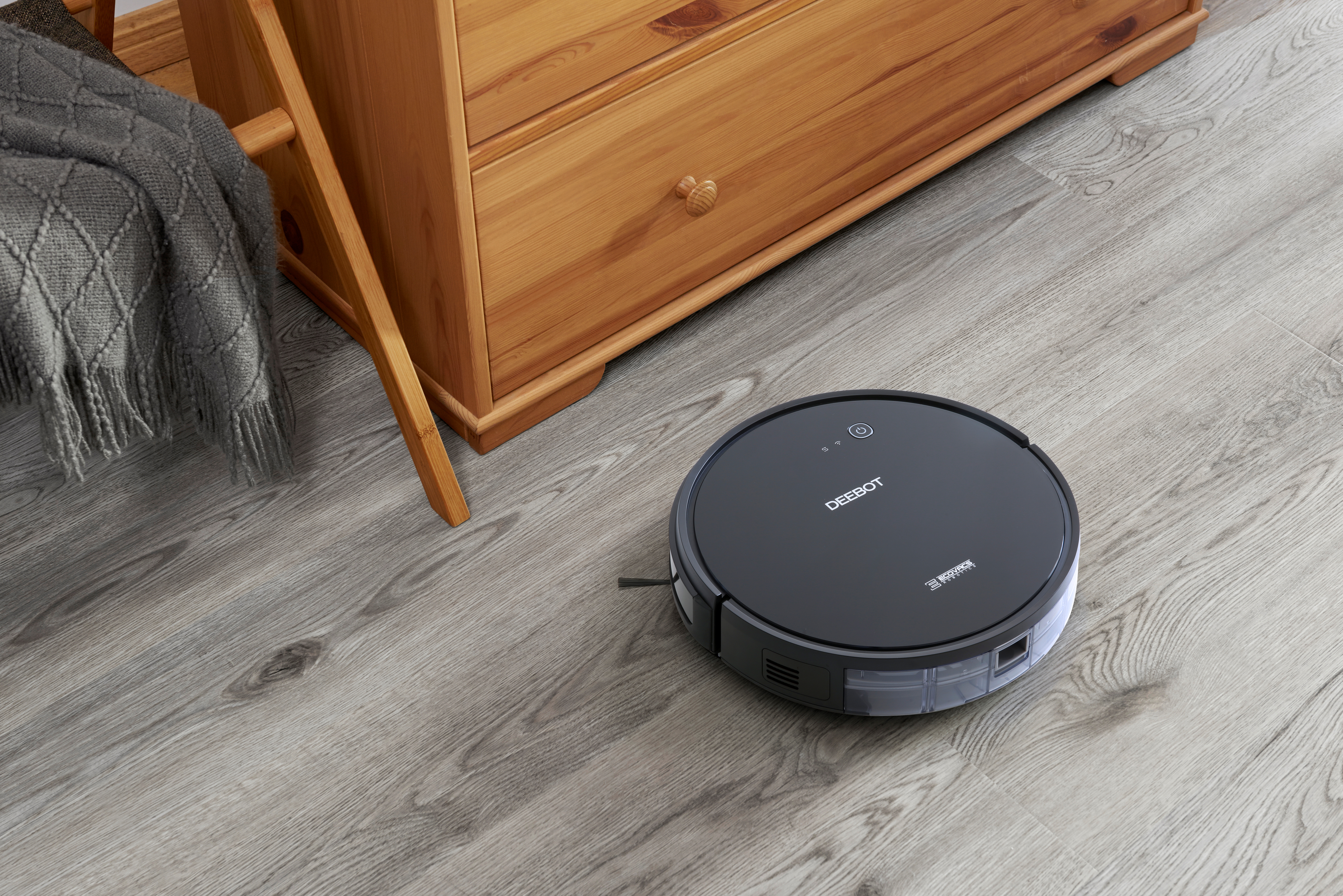 Ein schwarzer Roboter-Staubsauger auf einem grauen, holzähnlichen Boden. Eine Holzkommode im Hintergrund.
