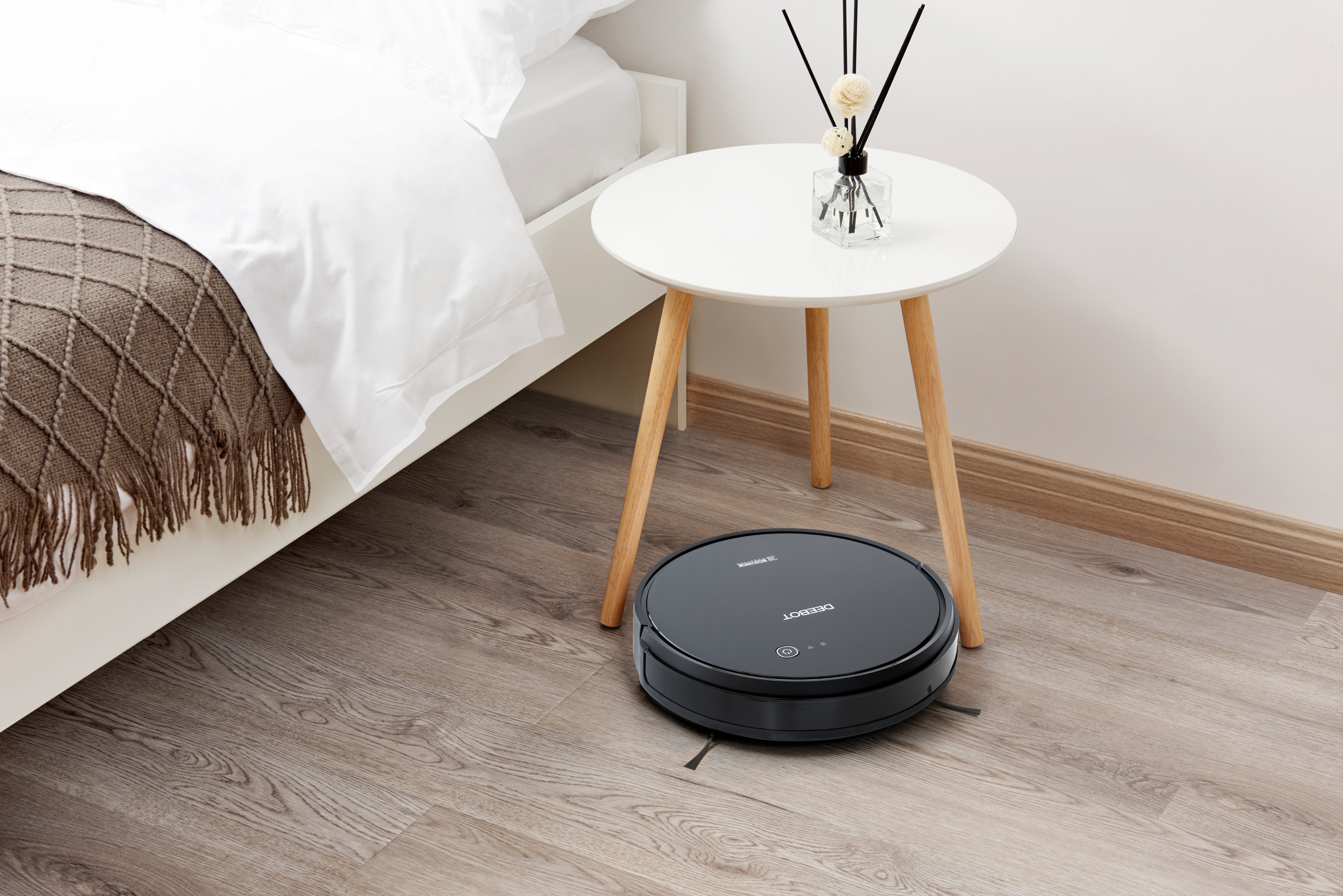 Roboter-Staubsauger auf dem Boden. Bett und Beistelltisch mit Blumenvase.