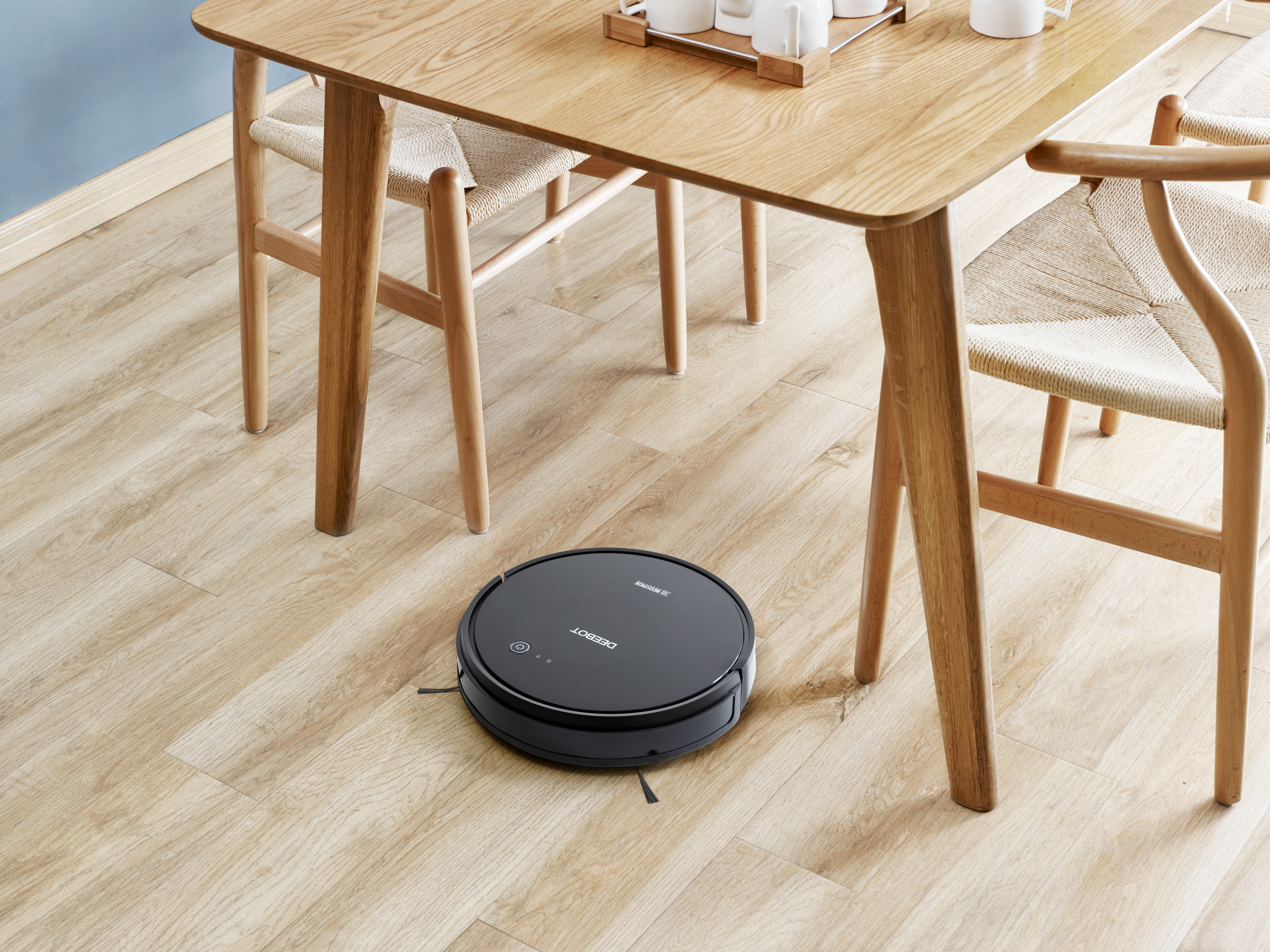 Ein schwarzer Roboter-Staubsauger auf hellem Holzboden, in der Nähe von Tisch und Stühlen.