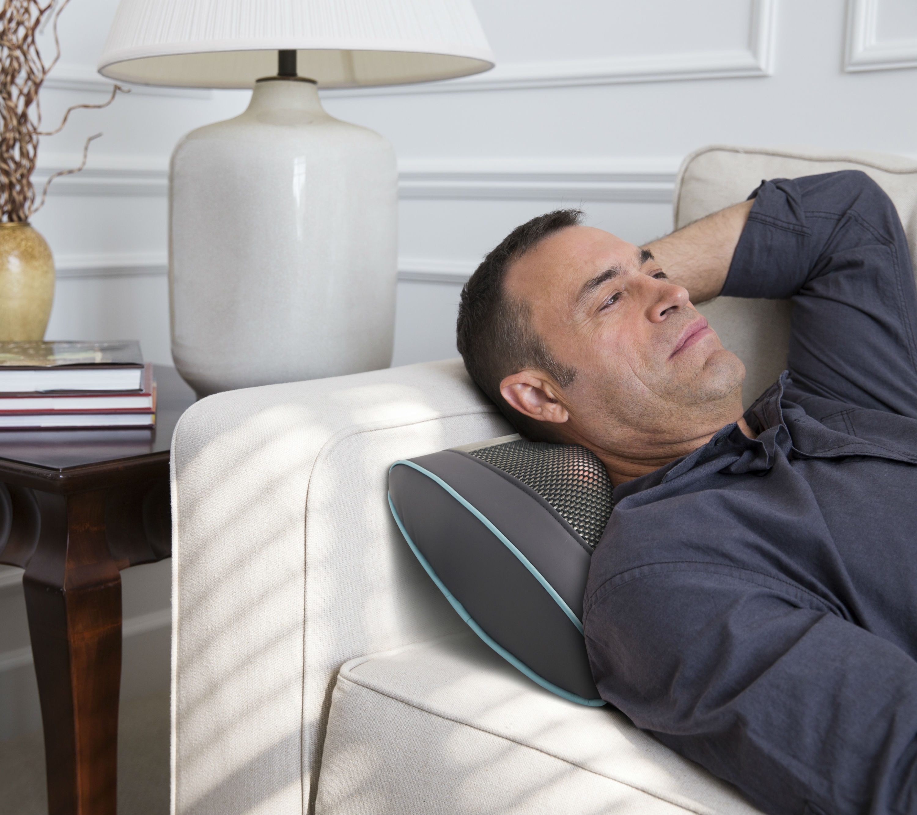 Mann entspannt sich auf einem Sofa mit einem Nackenmassagegerät. Eine Lampe und Bücher befinden sich links.