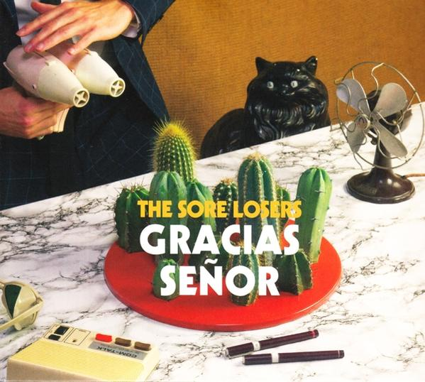Cactussen op rode plaat. Persoon met haardrogers. Zwarte kattenstandbeeld en ventilator. Tekst: Gracias Señor, The Sore Losers.