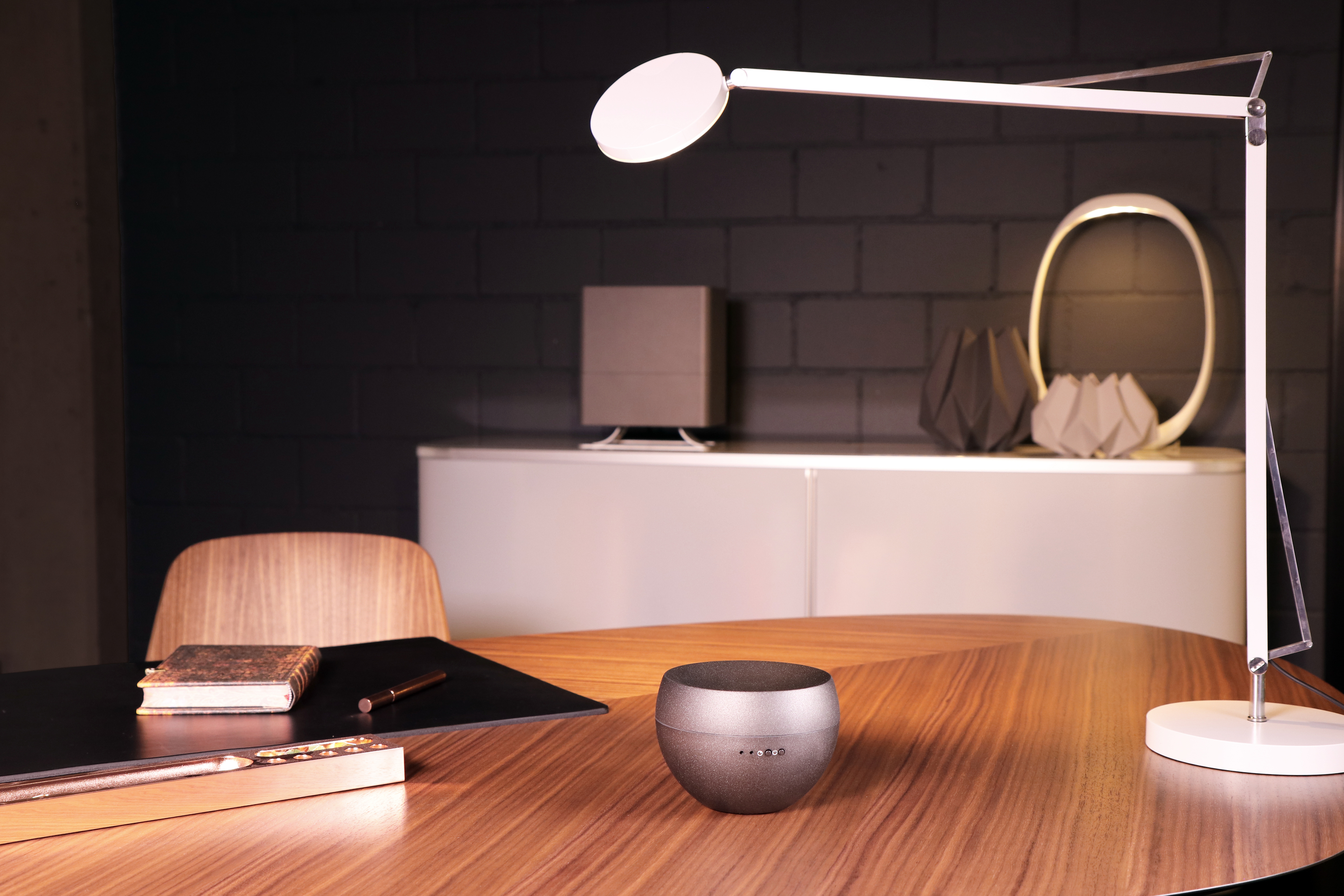 Schreibtischlampe beleuchtet einen Schreibtisch mit Lautsprecher. Notizbuch und Stift liegen auf dem Tisch. Hintergrund hat Deko.