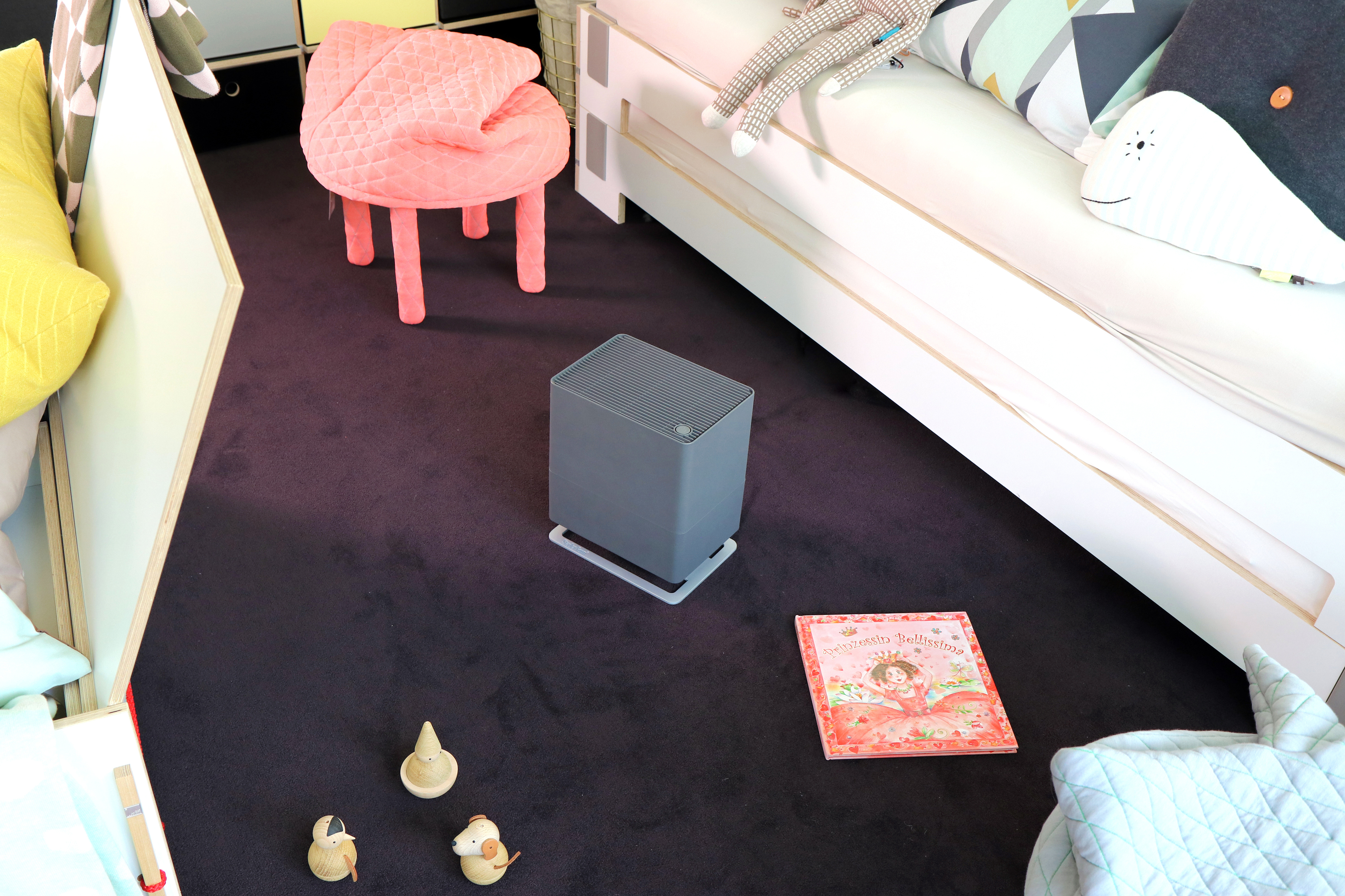 Ein Kinderzimmer mit Bett, rosa Stuhl und einem grauen Luftreiniger auf dem Boden. Ein Buch liegt am Boden.