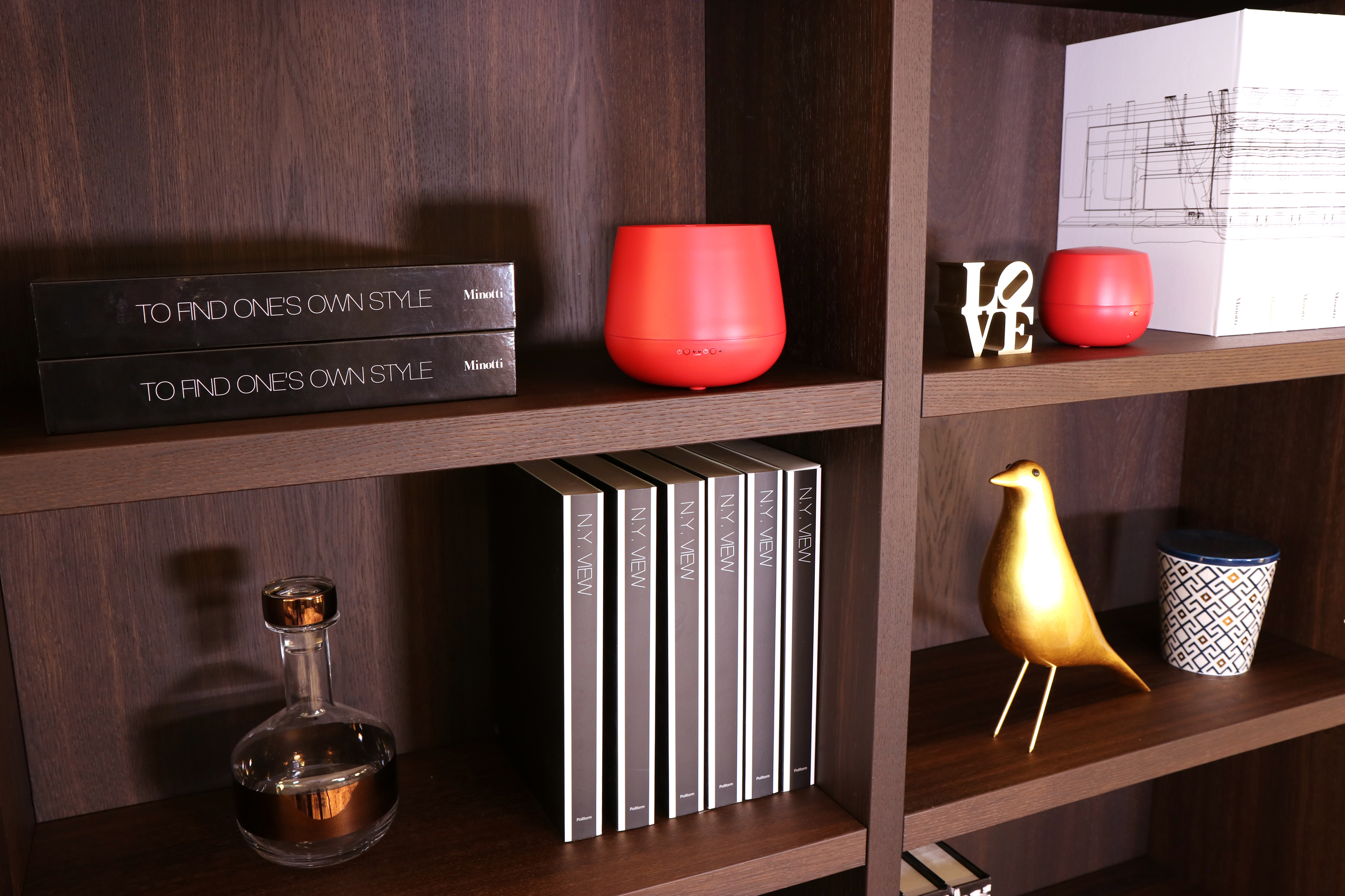 Ein Holzregal mit verschiedenen Gegenständen. Enthält Bücher, einen roten Diffusor, einen goldenen Vogel und Dekorationsartikel.