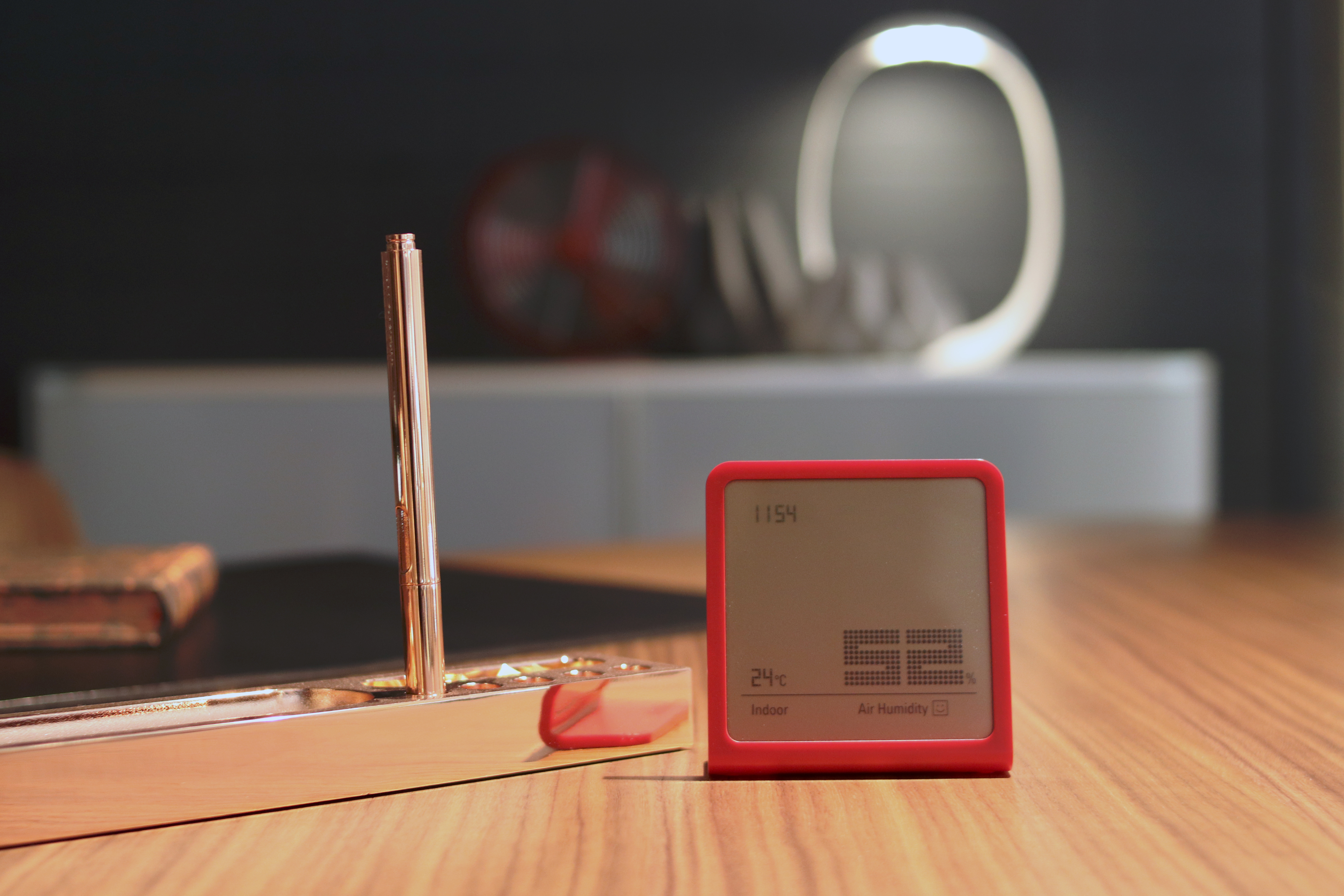 Schreibtisch mit rotem Digitalthermometer, Stifthalter, Lampe und Buch. Holzoberfläche.