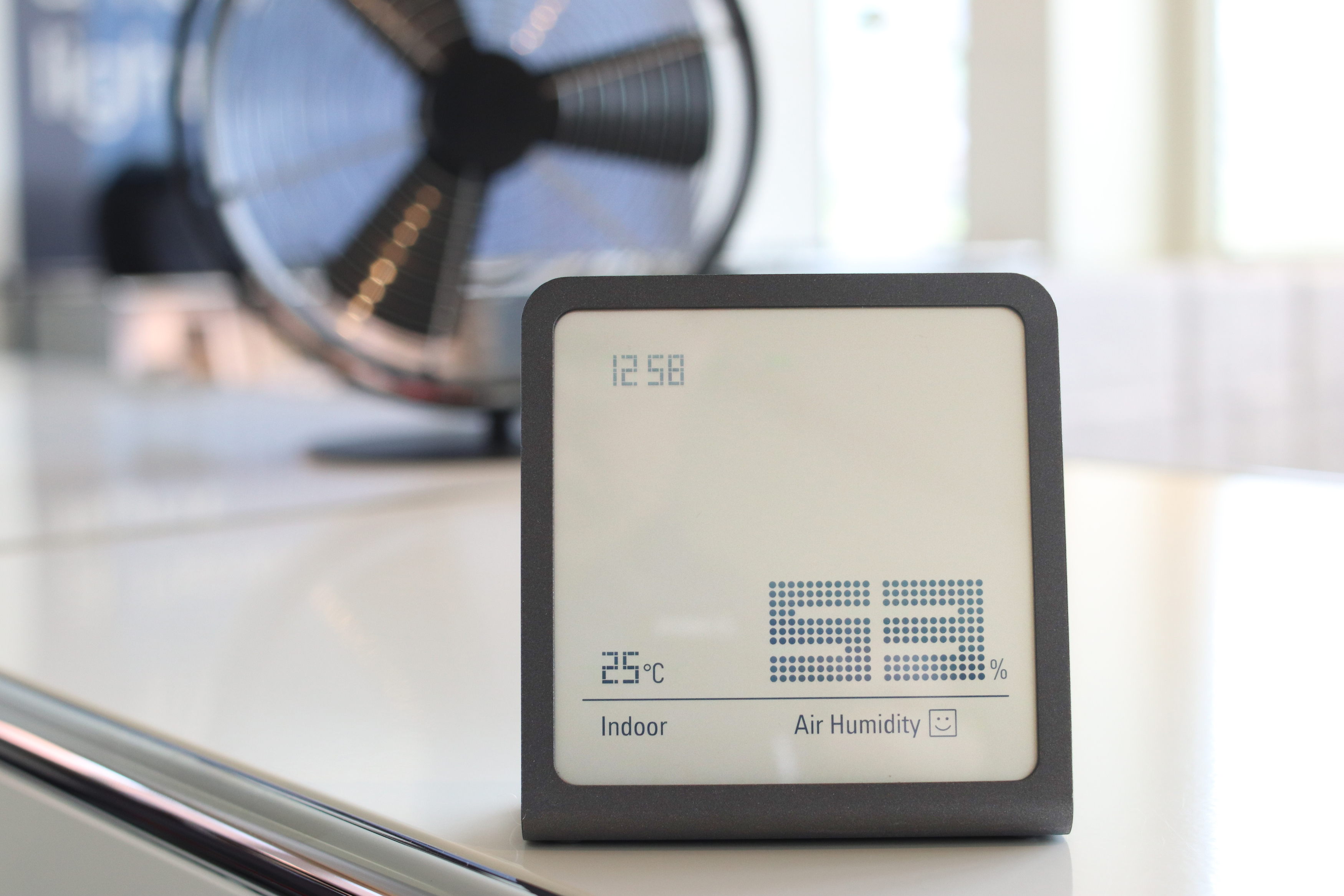 Graues Thermometer mit Digitalanzeige, die Zeit, Temperatur und Luftfeuchtigkeit anzeigt. Weiße Oberfläche.
