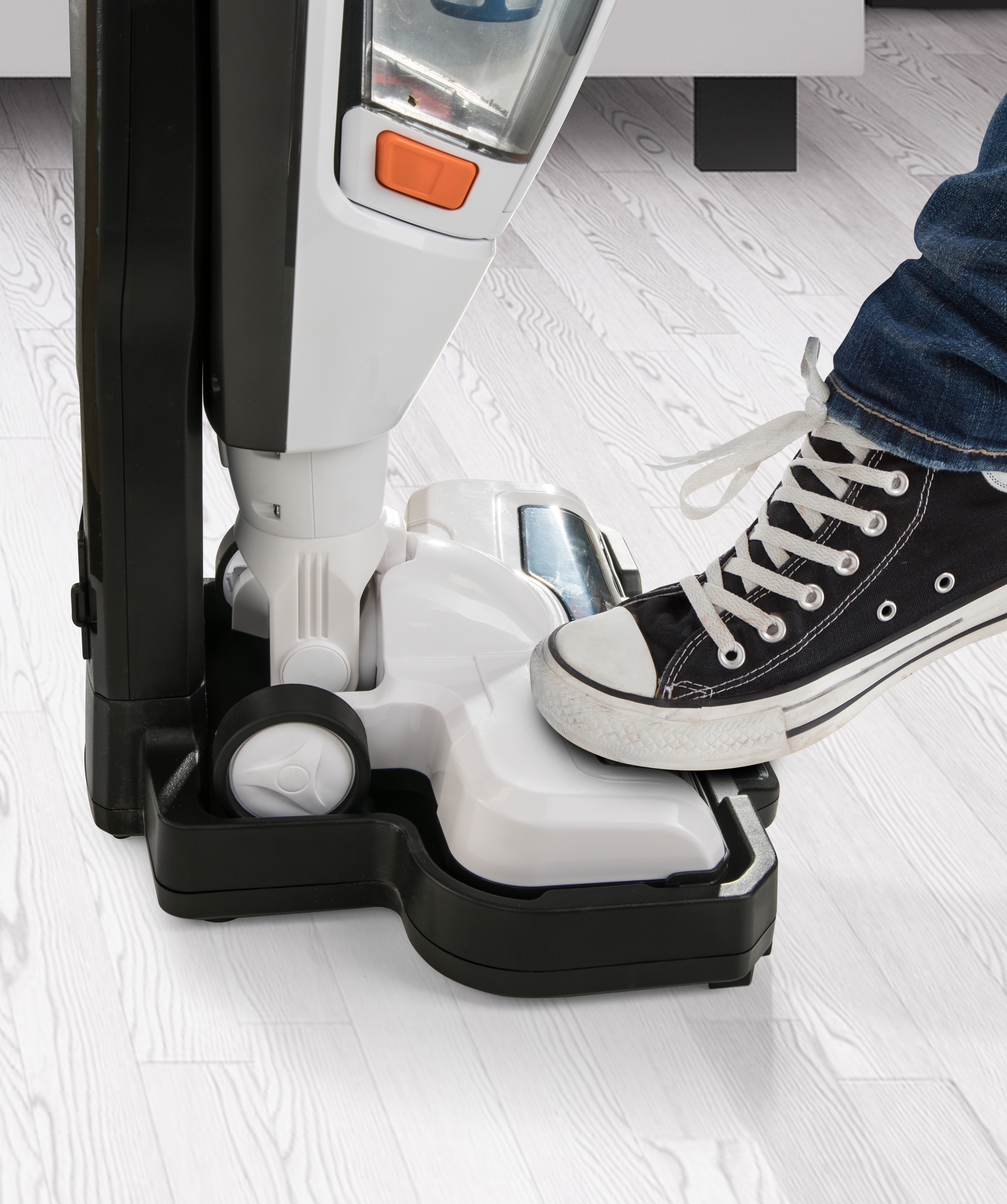 Fuß tritt auf einen weiß-schwarzen Staubsauger. Person trägt schwarze Schuhe.