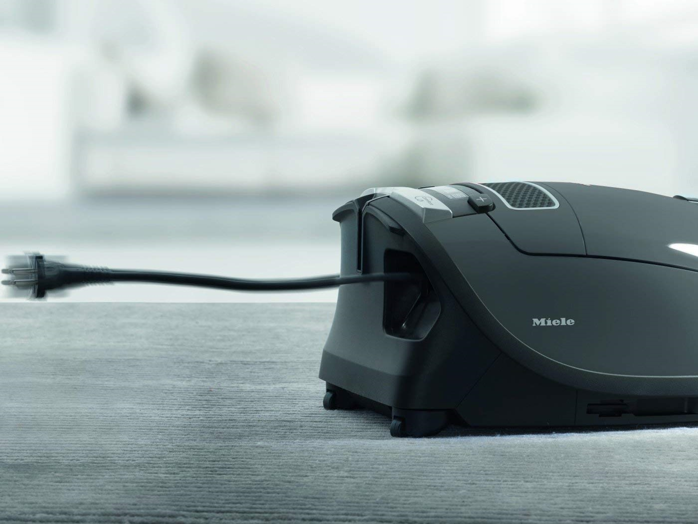 Schwarzer Miele Staubsauger mit eingestecktem Kabel. Der Staubsauger steht auf einer hellen Oberfläche.