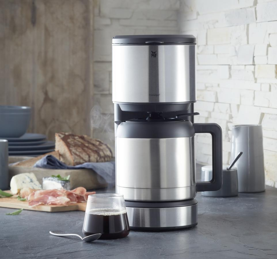 Cafetera de acero inoxidable con jarra de cristal, alimentos en la mesa.