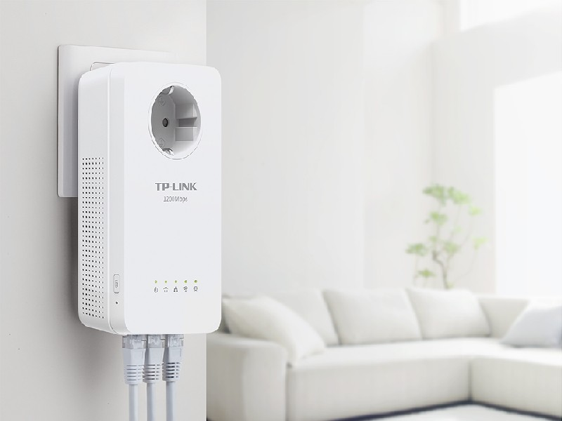 Dispositivo TP-Link blanco enchufado a la pared, con cables ethernet. Sofá y planta detrás.