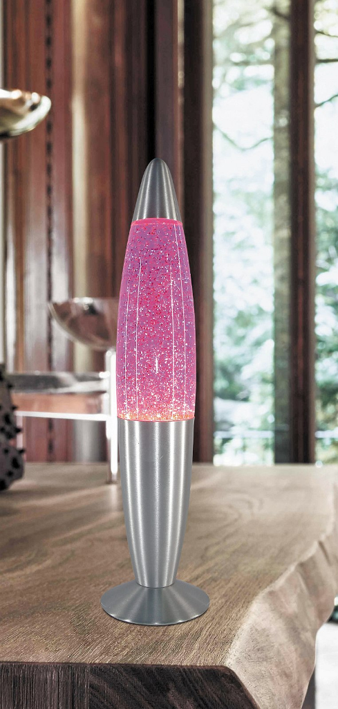 Egy ezüst lávalámpa rózsaszín csillámmal egy faasztalon áll. A háttérben egy ablak fák. A lámpa be van kapcsolva.
