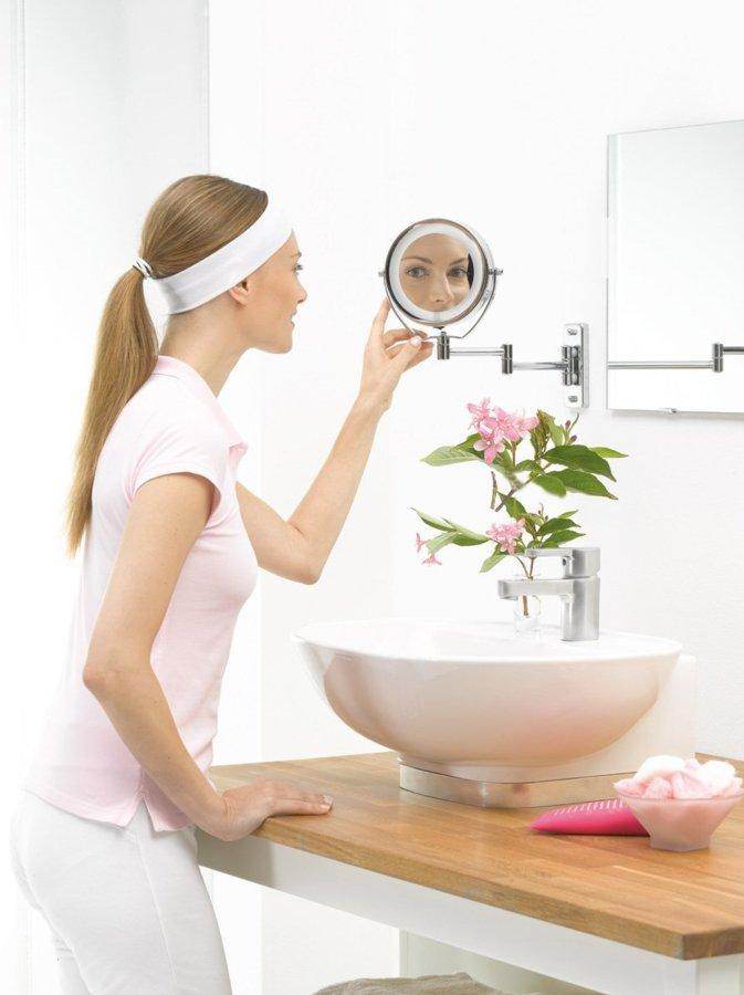 Una mujer con una camisa rosa mira un espejo de aumento sobre un lavabo con flores.