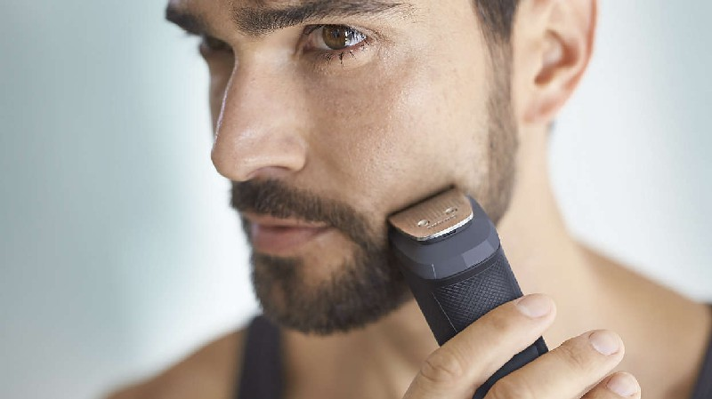 Un hombre se afeita la barba con una recortadora negra. El fondo está borroso.
