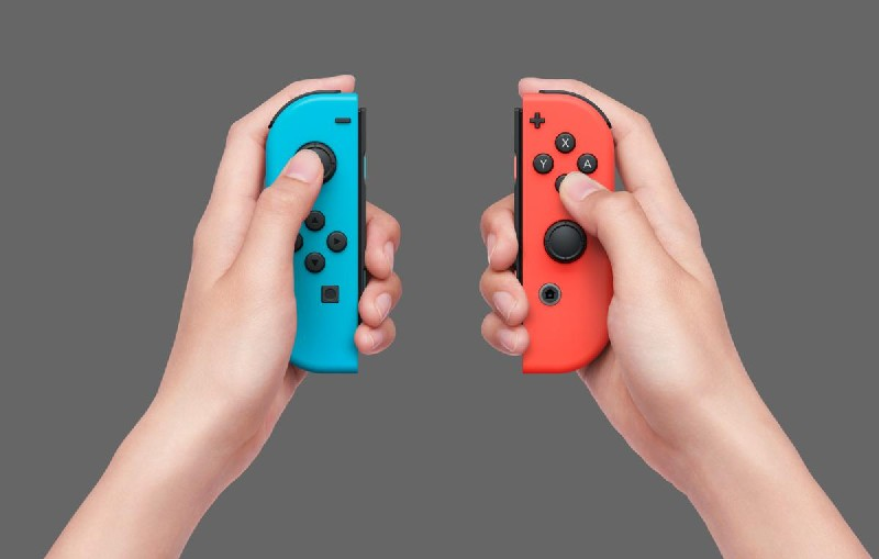 Dos manos sosteniendo Joy-Cons azul y rojo de Nintendo Switch sobre un fondo gris.